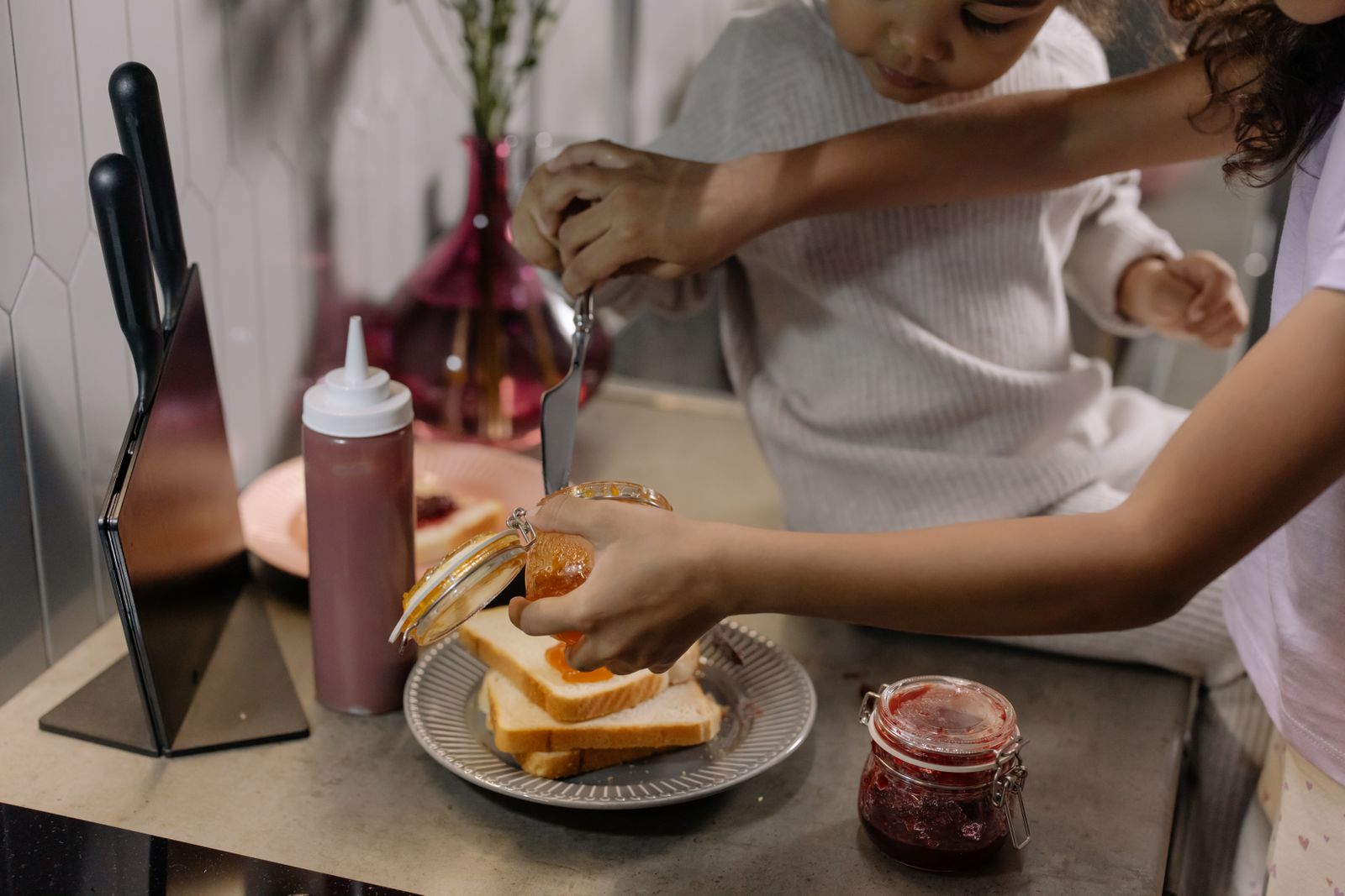 A mother and daughter bond while making a sandwich with jam on a kitchen counter.